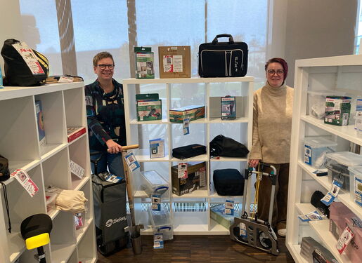 Das Bild zeigt zwei Personen, die vor Regalen mit verschiedenen Produkten stehen. Die Regale enthalten Artikel wie Taschen, Kisten und andere verpackte Waren.