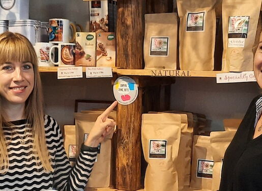 Das Bild zeigt zwei Personen, die vor Regalen mit verschiedenen verpackten Produkten stehen. Eine Person zeigt auf einen runden Aufkleber am Regal.