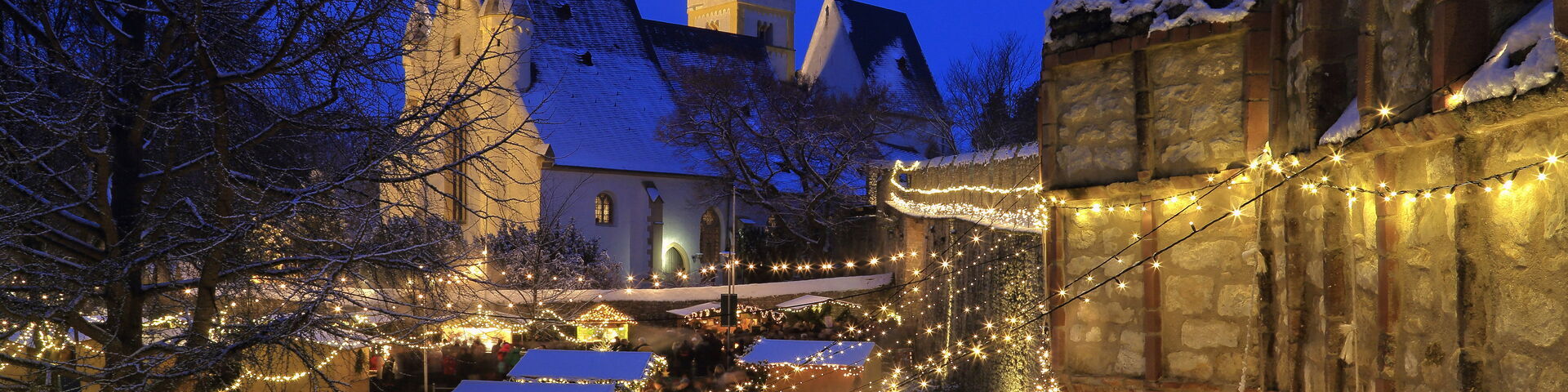 Das Bild zeigt eine festliche Weihnachtsmarktszene am Abend mit zahlreichen Lichterketten, die den Bereich beleuchten. Es gibt mehrere kleine Holzstände, die mit Lichtern und Schnee dekoriert sind, und eine Menschenmenge, die zwischen i