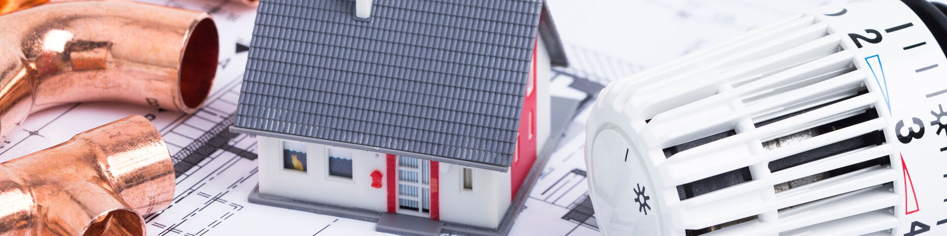 Auf dem Bild ist ein Bauplan zu sehen. Darauf ist ein kleines Häuschen, eine Zange, Rohre und ein Heizungsthermostat platziert.
