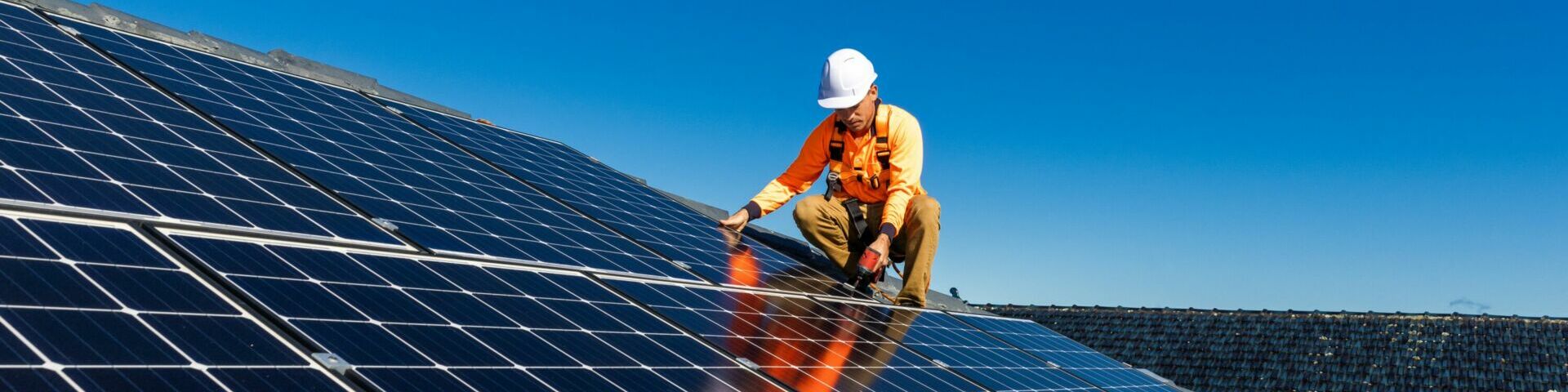 Das Bild zeigt eine Person, die eine orangefarbene Sicherheitsweste und einen weißen Schutzhelm trägt und auf einem Dach Solarpaneele installiert oder wartet. Die Person kniet und scheint eines der Solarpaneele zu justieren.