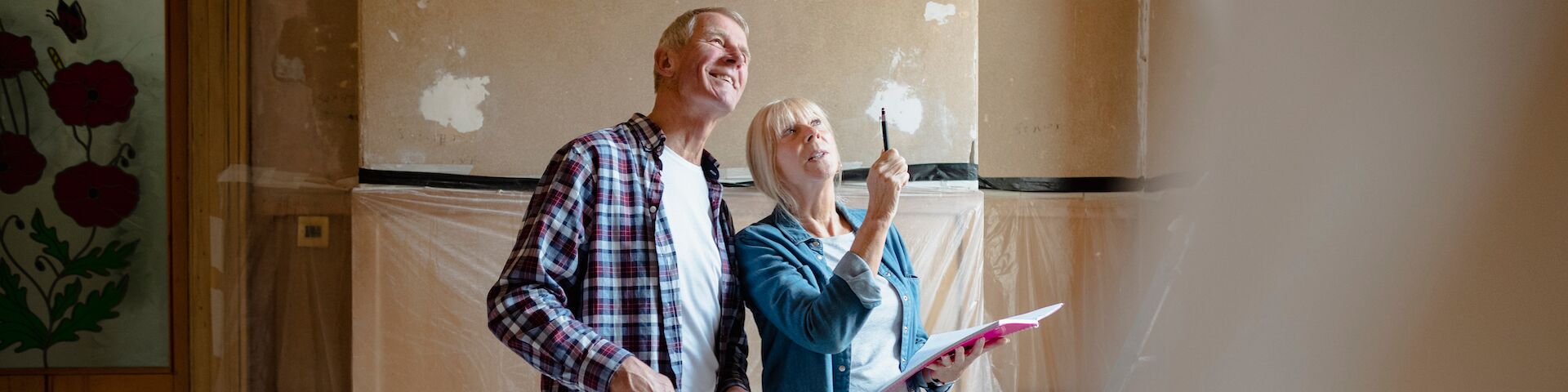 Eine Frau und ein Mann stehen in einem Raum, der renoviert wird. Die Frau hält ein Notizbuch und zeigt auf etwas an der Wand, während der Mann in die gleiche Richtung schaut. Die Wände haben Putzstellen.