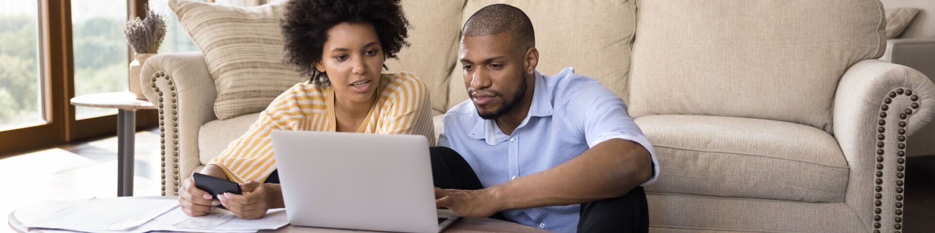 Das Bild zeigt zwei Personen, die auf dem Boden vor einem Couchtisch sitzen. Vor ihnen steht ein aufgeklappter Laptop. Eine Person hält einen Taschenrechner und einige Papiere, während die andere auf den Laptop-Bildschirm zeigt.