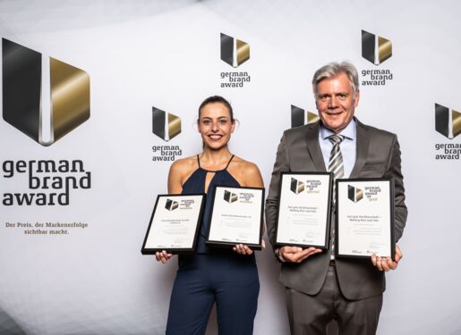 Verena Schulze und Peter Hausen zeigen gerahmte Zertifikate vor einem Hintergrund, auf dem "German Brand Award" steht.  Jede Person hält zwei Zertifikate, sodass insgesamt vier Zertifikate im Bild zu sehen sind.