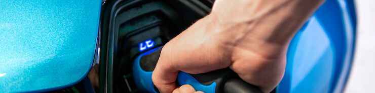 es wird ein blaues E-Auto betankt. Eine Hand hält das Ladekabel und steckt diese ins Auto.