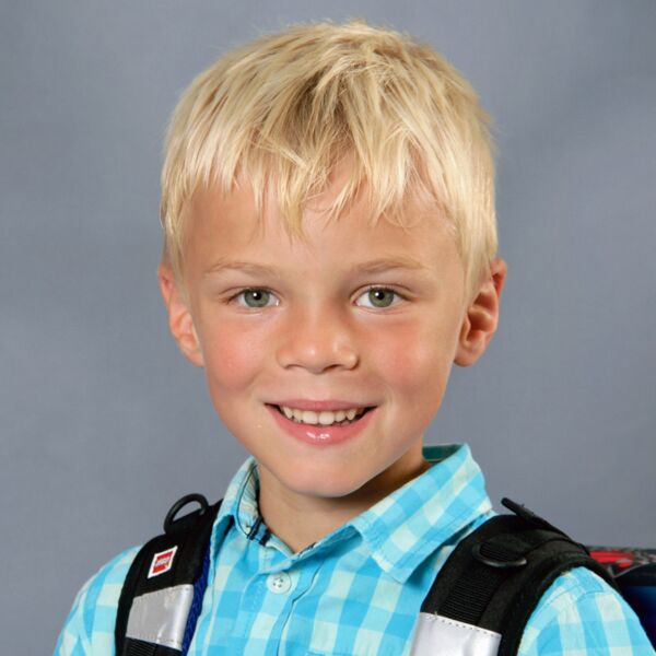 Schulkind aus Weiler bei Bingen. Das Bild zeigt ein Kind mit blondem Haar, das ein blau-weiß kariertes Hemd trägt. Das Kind hat außerdem einen schwarzen Rucksack mit reflektierenden Riemen und Schnallen. Der Hintergrund ist einfarbig grau.
