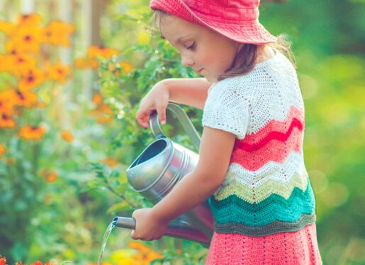Das Bild zeigt ein kleines Mädchen, das einen roten Hut und ein buntes Kleid trägt und mit einer orangefarbenen Gießkanne Pflanzen in einem Garten gießt. Der Garten ist mit verschiedenen Blumen gefüllt, darunter orange und weiße Blüten.