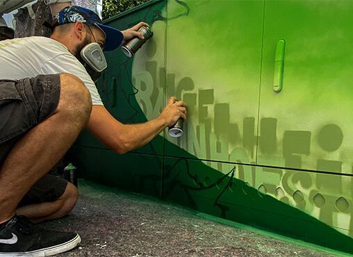 Das Bild zeigt einen Mann, der Graffiti auf eine grüne Wand sprüht. Der Mann hockt und trägt legere Kleidung. Das Graffiti ist in Grüntönen gehalten und besteht aus Buchstaben. Ingelheim Bahnhof ist zu lesen.