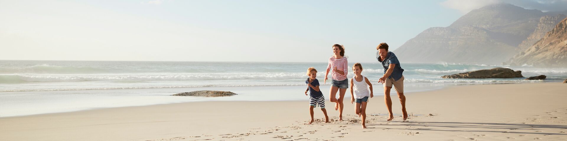 Eine Familie mit vier Personen, die am Strand entlanglaufen. Im Hintergrund sieht man das Meer mit sanften Wellen und entfernten Bergen unter einem klaren Himmel.