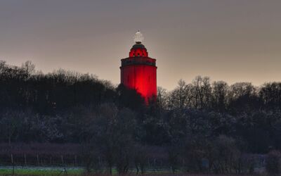 Ingelummer Kerz in Ingelheim bei Dämmerung. Das Bild zeigt einen hohen, zylindrischen Turm mit einem Licht an der Spitze, das rot leuchtet. Der Turm ist von Bäumen und Vegetation umgeben.