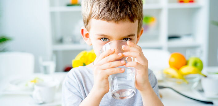 Das Bild zeigt einen Jungen, der an einem Tisch sitzt und aus einem Glas Wasser trinkt. Im Hintergrund sind unscharf verschiedene Gegenstände wie Obst (Orangen, Bananen, Äpfel) und Geschirr zu sehen.