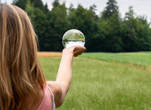 Eine Frau mit langen Haaren steht in einem grasbewachsenen Feld und hält ein klares, kugelförmiges Objekt auf Armlänge. Im Hintergrund sind Bäume und ein offener Himmel zu sehen.