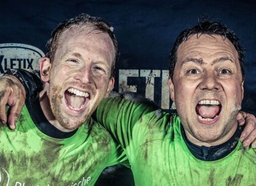 Jan Lubkowski und Viktor Hipp nach dem XLETIX Hindernislauf freudig umarmend. Beide sind mit Schlamm bedeckt.