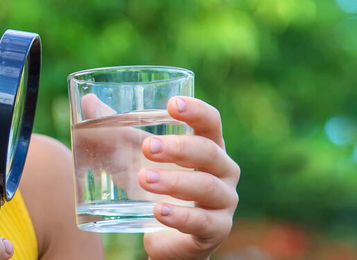 Das Bild zeigt ein Mädchen, das in einer Hand eine Lupe und in der anderen ein Glas Wasser hält. Sie schaut durch die Lupe ins Glas. Der Hintergrund ist unscharf und zeigt grünes Laub,