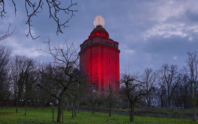 Ingelummer Kerz in Ingelheim am frühen Morgen. Das Bild zeigt einen hohen Turm, der mit roten Lichtern beleuchtet ist. Der Turm hat ein markantes architektonisches Design mit mehreren Ebenen und einer großen, leuchtenden Kugelstruktur.