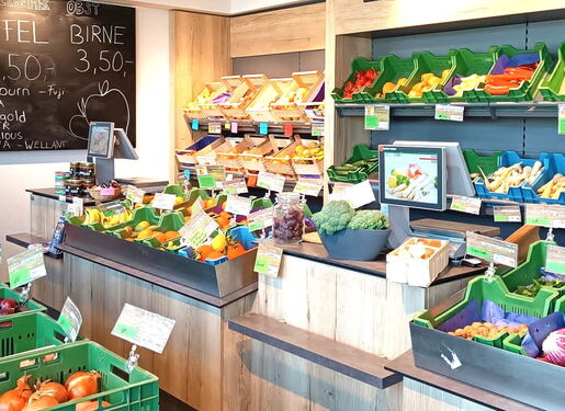 Das Bild zeigt das Innere eines Lebensmittelgeschäfts mit verschiedenen Obst- und Gemüsesorten. Es gibt grüne Plastikkisten, die mit verschiedenen Arten von Produkten wie Äpfeln, Zwiebeln und anderen Gemüsesorten gefüllt sind.
