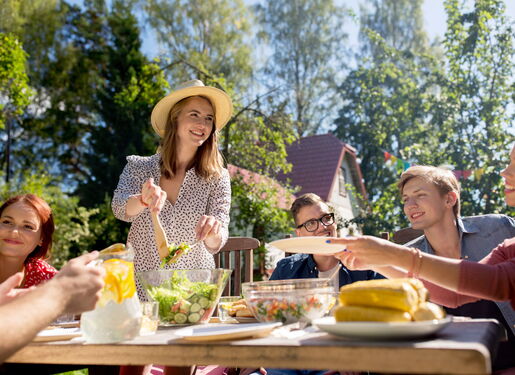 Zu sehen ist eine Familie draußen im Garten beim Sommergrillen. Es gibt Salate und Maiskolben. Im Hintergrund befindet sich ein Haus und Bäume.