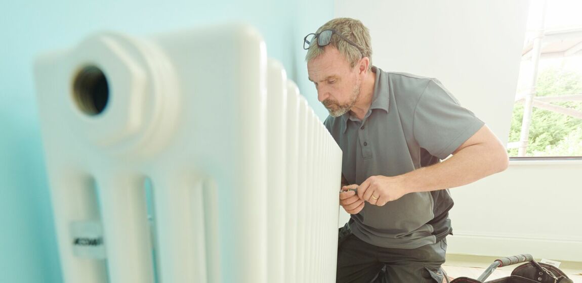 Das Bild zeigt eine Person, die auf einem mit einem Tuch bedeckten Boden kniet und an einem weißen Heizkörper arbeitet. Die Person benutzt Werkzeuge aus einer offenen Werkzeugtasche, die in der Nähe platziert ist.