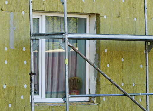 Das Bild zeigt einen Abschnitt eines Gebäudes, das sich in der Renovierung befindet. Die Außenwände sind mit Dämmmaterial bedeckt. Es sind zwei Fenster sichtbar, und vor dem Gebäude ist ein Gerüst aufgebaut.