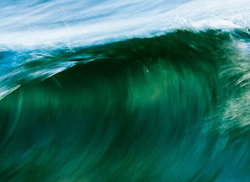 Das Bild zeigt eine große Ozeanwelle in verschiedenen Blau- und Grüntönen. Die Welle rollt sich über und erzeugt eine dynamische und kraftvolle Szene. Das Wasser scheint in Bewegung zu sein, und am Kamm der Welle bildet sich weißer Schaum.