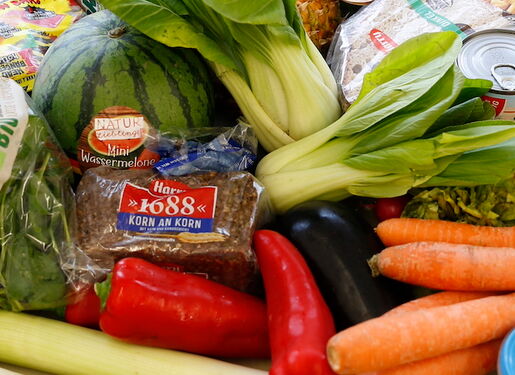 Das Bild zeigt eine Vielzahl frischer Gemüse, Früchte und anderer Lebensmittel, die auf einer Fläche ausgebreitet sind. Sichtbare Artikel umfassen Blumenkohl, Karotten, Pak Choi, rote Paprika, Bananen, Äpfel, Zitronen, Limetten und Blattgemüse.