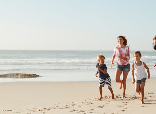 Eine Familie mit vier Personen, die am Strand entlanglaufen. Im Hintergrund sieht man das Meer mit sanften Wellen und entfernten Bergen unter einem klaren Himmel.
