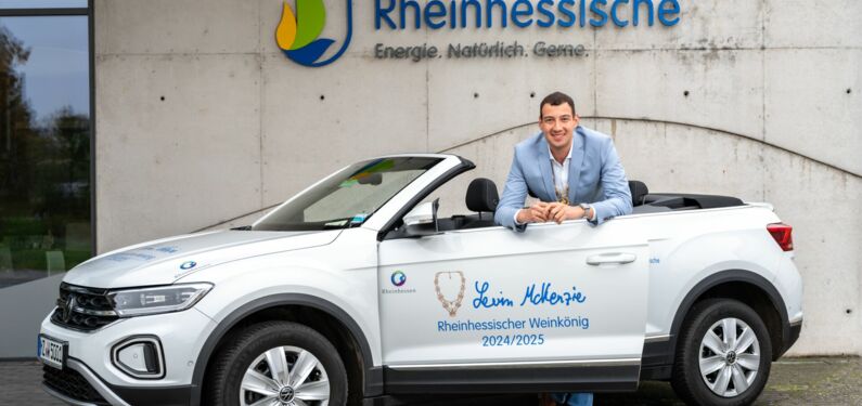 Das Bild zeigt Levin McKenzie, der sich an die Tür eines weißen Cabriolets lehnt. Das Auto hat Texte und Logos darauf, darunter "Rheinhessischer Weinkönig 2024/2025" und "Levin McKenzie." Im Hintergrund ist Kundencenter der  "Rheinhessischen" zu sehen.