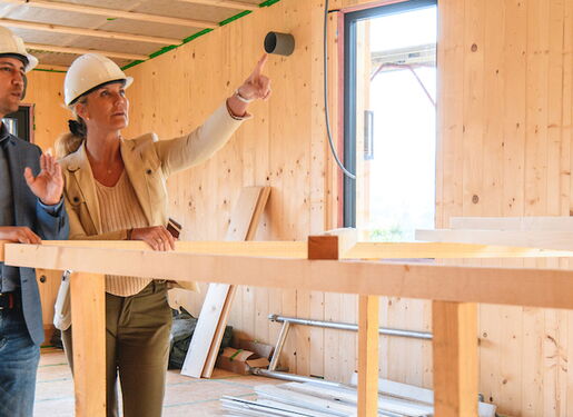 Zwei Personen, die Schutzhelme tragen und sich in einer Holzkonstruktion befinden, die im Bau ist. Um sie herum sind Holzbalken und Baumaterialien sichtbar.