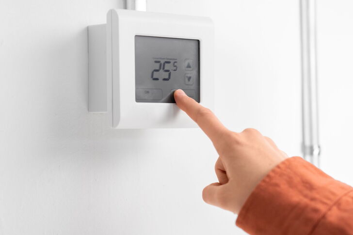 Das Bild zeigt ein digitales Thermostat, das an einer Wand montiert ist. Eine Hand berührt den Bildschirm und drückt speziell den Pfeil nach oben. Das Display des Thermostats zeigt "25°C" an.