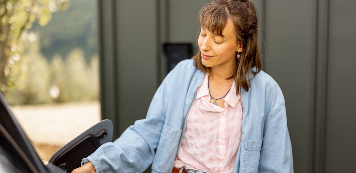 Das Motiv zeigt eine junge Frau in einer blauen Jeanskombi. Sie steht neben Ihrem E-mobil lädt Ihr Fahrzeug an einer Wallbox.