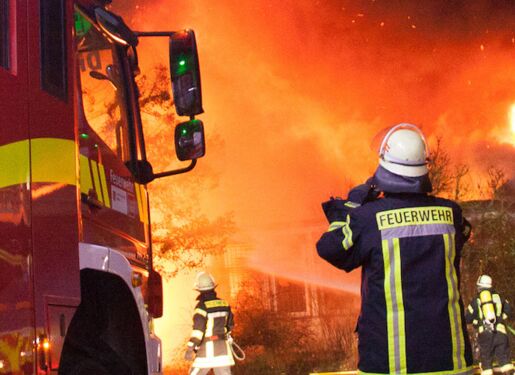 Eine Gruppe von Feuerwehrleuten, die aktiv daran arbeiten, ein großes Feuer zu löschen. Es sind zwei Feuerwehrautos mit der Aufschrift "FEUERWEHR MAINZ" zu sehen. Die Feuerwehrleute tragen Schutzkleidung, einschließlich Helmen und reflektierenden Jacken.