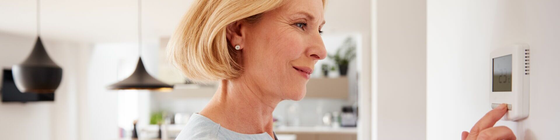 Eine Frau mit kurzen blonden Haaren, die ein hellblaues Hemd trägt und einen Thermostat an der Wand einstellt. Im Hintergrund sieht man eine moderne Küche und einen Essbereich mit Holzmöbeln und schwarzen Pendelleuchten.