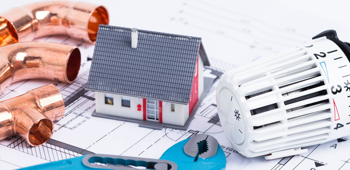 Auf dem Bild ist ein Bauplan zu sehen. Darauf ist ein kleines Häuschen, eine Zange, Rohre und ein Heizungsthermostat platziert.