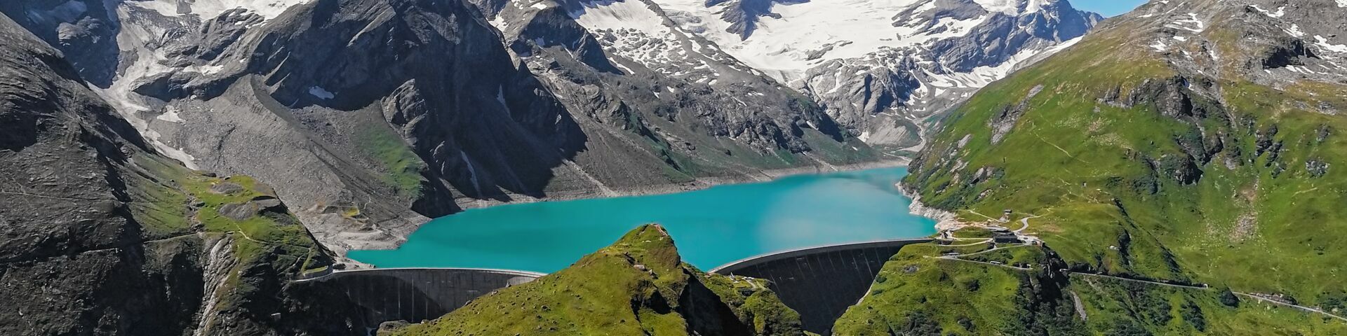 Das Bild zeigt eine malerische Landschaft mit schneebedeckten Berggipfeln im Hintergrund. In der Mitte des Bildes befindet sich ein türkisfarbener See, der von steilen Bergen umgeben ist. Der See scheint von einem Damm gehalten zu werden.