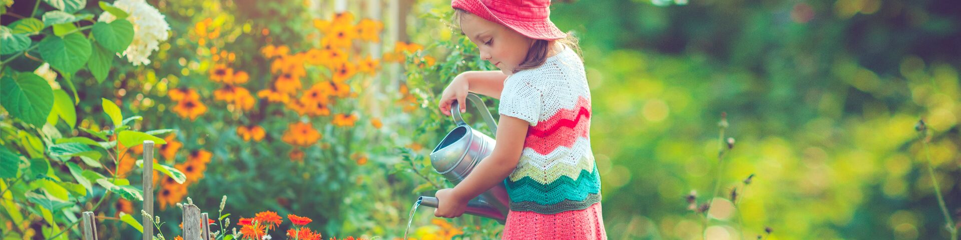 Das Bild zeigt ein kleines Mädchen, das einen roten Hut und ein buntes Kleid trägt und mit einer orangefarbenen Gießkanne Pflanzen in einem Garten gießt. Der Garten ist mit verschiedenen Blumen gefüllt, darunter orange und weiße Blüten.