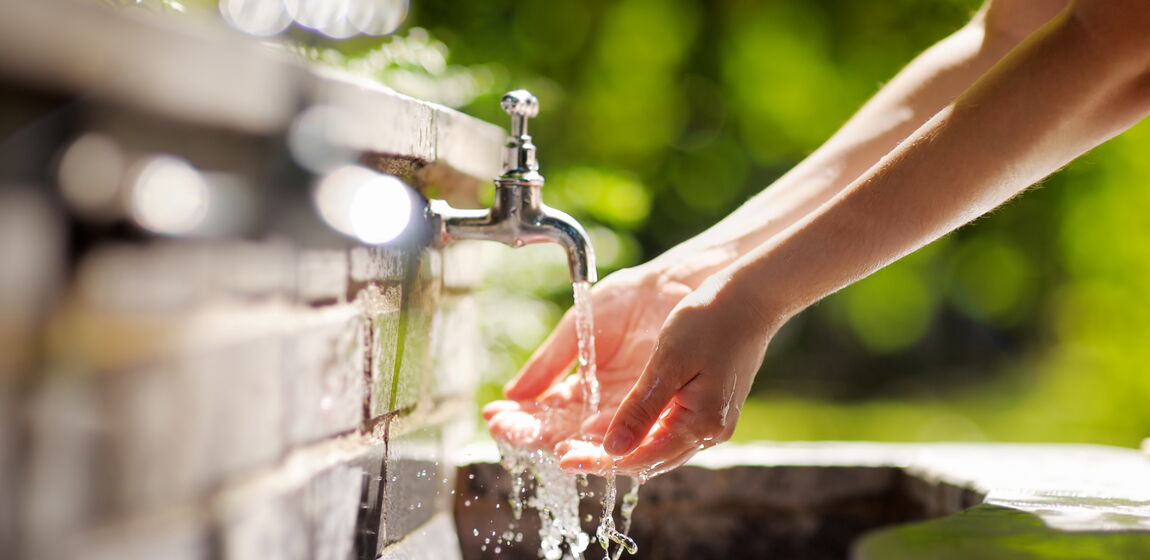 Das Bild zeigt eine Person, die sich unter einem Wasserhahn an einer Steinmauer die Hände wäscht. Das Wasser fließt in ein Becken darunter. Die Szene wirkt natürlich und alltäglich.