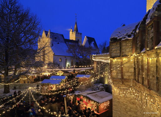 Das Bild zeigt eine festliche Weihnachtsmarktszene am Abend mit zahlreichen Lichterketten, die den Bereich beleuchten. Es gibt mehrere kleine Holzstände, die mit Lichtern und Schnee dekoriert sind, und eine Menschenmenge, die zwischen i