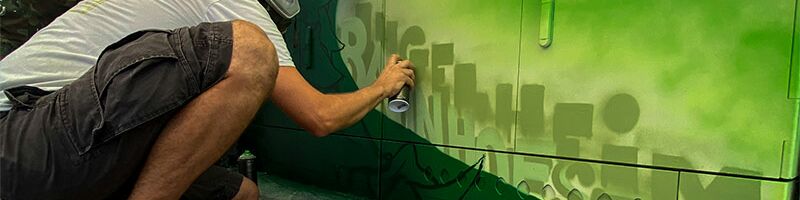 Das Bild zeigt einen Mann, der Graffiti auf eine grüne Wand sprüht. Der Mann hockt und trägt legere Kleidung. Das Graffiti ist in Grüntönen gehalten und besteht aus Buchstaben. Ingelheim Bahnhof ist zu lesen.