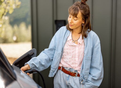 Das Motiv zeigt eine junge Frau in einer blauen Jeanskombi. Sie steht neben Ihrem E-mobil lädt Ihr Fahrzeug an einer Wallbox.