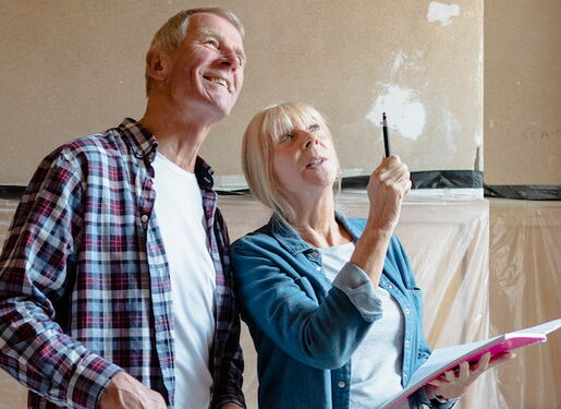 Eine Frau und ein Mann stehen in einem Raum, der renoviert wird. Die Frau hält ein Notizbuch und zeigt auf etwas an der Wand, während der Mann in die gleiche Richtung schaut. Die Wände haben Putzstellen.