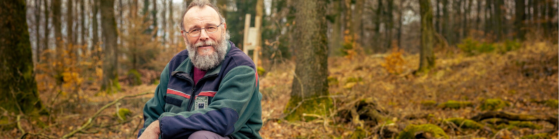Ein Förster sitzt auf einem gefällten Baumstamm im herbstlichen Wald. Er trägt grüne Jacke, lila Hose und ruht sich aus oder beobachtet die Umgebung zwischen kahlen Bäumen und Laub.