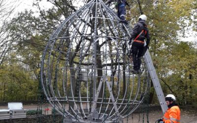 Das Bild zeigt drei Arbeiter, die Sicherheitsausrüstung und Helme tragen und eine große, metallene, tropfenförmige Struktur, die Spitze der Ingelummer Kerz in einer Außenumgebung zusammenbauen.