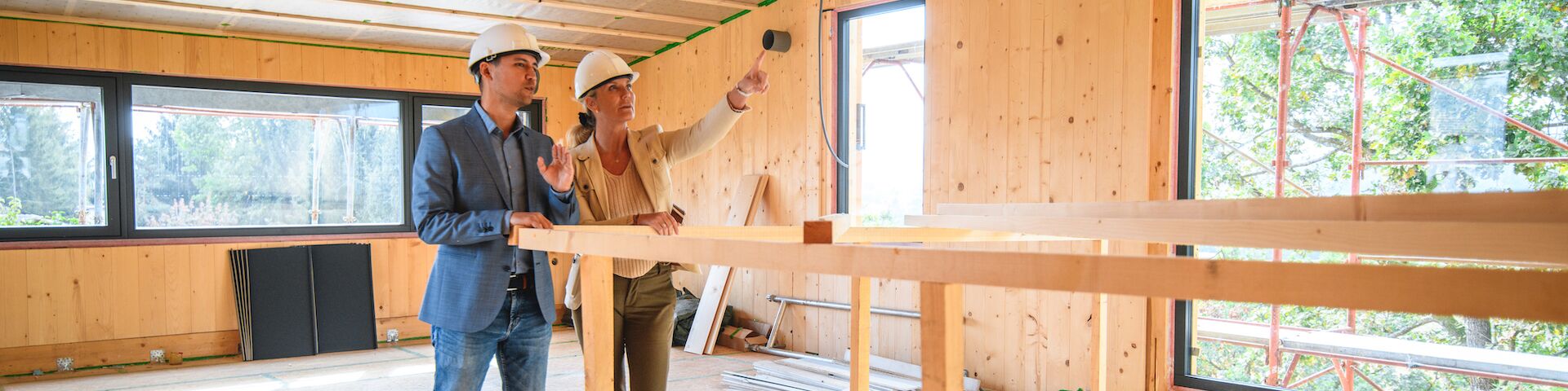 Zwei Personen, die Schutzhelme tragen und sich in einer Holzkonstruktion befinden, die im Bau ist. Um sie herum sind Holzbalken und Baumaterialien sichtbar.
