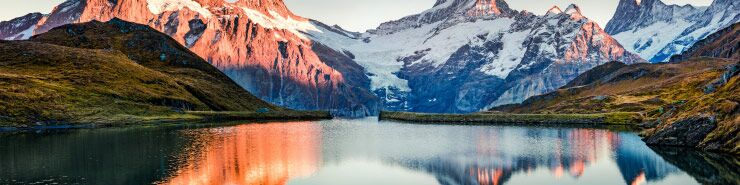 Das Bild zeigt eine ruhige Berglandschaft mit einem stillen See im Vordergrund. Der See spiegelt die umliegenden schneebedeckten Berge und den klaren Himmel wider. Die Berge sind vom warmen Licht des Sonnenaufgangs beleuchtet.