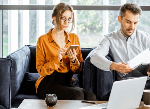 Eine Frau und ein Mann sitzen auf einem dunkelblauen Sofa in einem Büro. Die Frau trägt ein orangefarbenes Hemd und tippt auf einem Smartphone, während der Mann einige Papiere in der Hand hält.