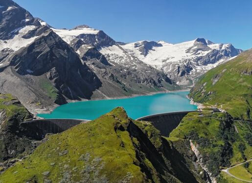 Das Bild zeigt eine malerische Landschaft mit schneebedeckten Berggipfeln im Hintergrund. In der Mitte des Bildes befindet sich ein türkisfarbener See, der von steilen Bergen umgeben ist. Der See scheint von einem Damm gehalten zu werden.