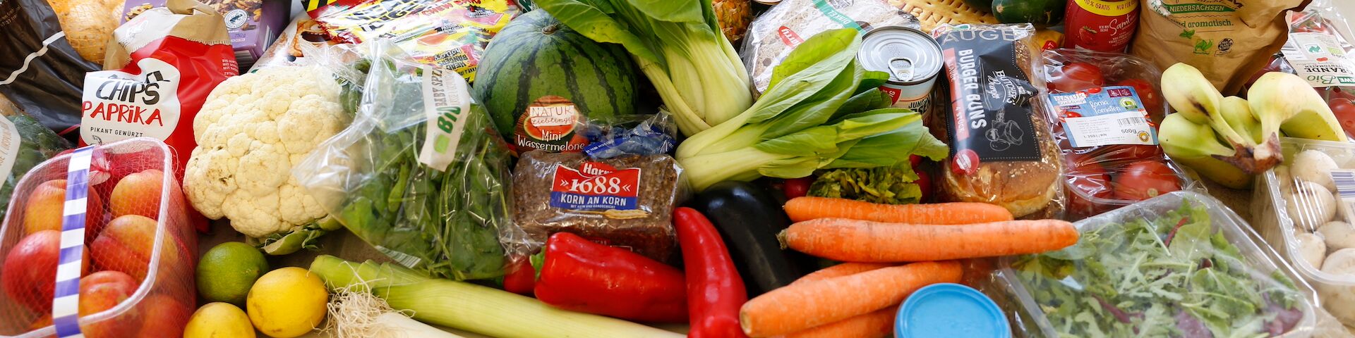Das Bild zeigt eine Vielzahl frischer Gemüse, Früchte und anderer Lebensmittel, die auf einer Fläche ausgebreitet sind. Sichtbare Artikel umfassen Blumenkohl, Karotten, Pak Choi, rote Paprika, Bananen, Äpfel, Zitronen, Limetten und Blattgemüse.