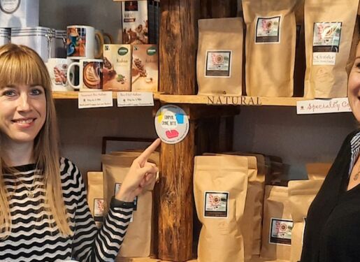 Das Bild zeigt zwei Personen, die vor Regalen mit verschiedenen verpackten Produkten stehen. Eine Person zeigt auf einen runden Aufkleber am Regal.