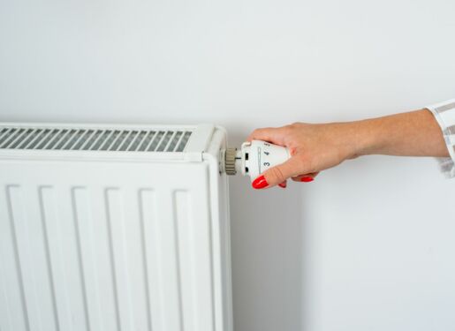Das Bild zeigt eine Hand, die den Thermostat an einem weißen Heizkörper einstellt. Die Hand hat rot lackierte Fingernägel und dreht den Knopf an der Seite des Heizkörpers, der Zahlen und Symbole zur Anzeige der verschiedenen Temperatureinstellungen hat.