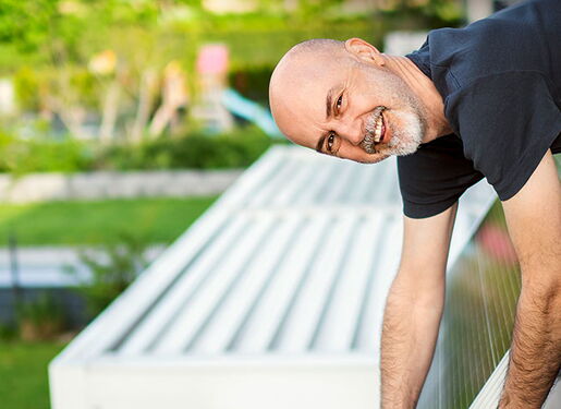 Das Bild zeigt eine Person, die sich über das Geländer eines Balkons lehnt und lächelt und an einem Solarpannel arbeitet.